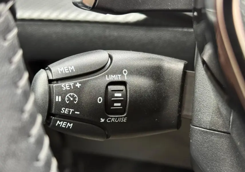 Gros plan sur la commande de régulateur de vitesse noire à gauche du volant dans une Peugeot 208 noire Perla Nera 2025.