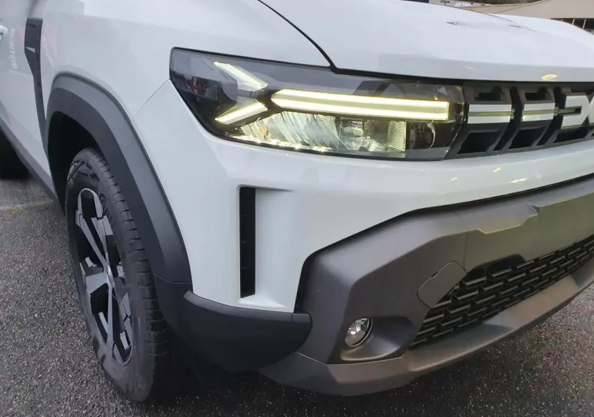 Gros plan sur l’avant gauche du Dacia Duster blanc glacier 2025, mettant en valeur les phares LED et la calandre noire.