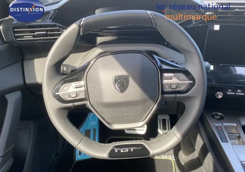 Vue rapprochée du volant cuir perforé noir avec badge GT du Peugeot 308 Hybrid 145ch, tableau de bord moderne en arrière-plan.