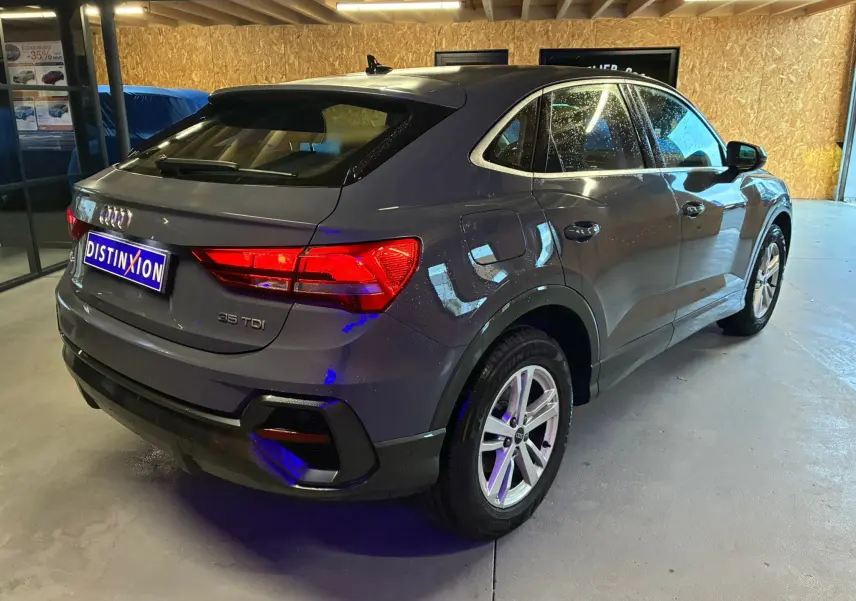 Audi Q3 Sportback gris en vue 3/4 arrière droit, avec jantes aluminium 17 pouces et feux arrière allumés.