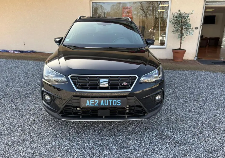 Vue frontale d'un SEAT Arona noir 2020 avec badge FR et calandre hexagonale, stationné sur un sol gravillonné devant un bâtiment.