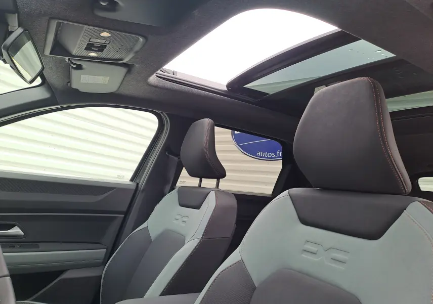 Vue intérieure avant du Dacia Bigster 2025 montrant les sièges gris et noir avec surpiqûres orange et toit ouvrant panoramique.