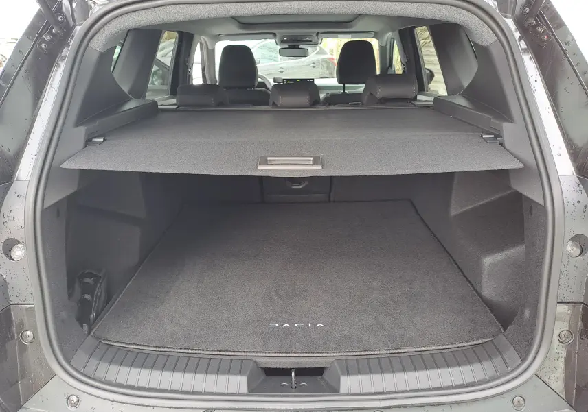 Coffre ouvert vu de l'arrière d'un Dacia Bigster noir nacré, avec cache-bagages et tapis de sol marqué DACIA.