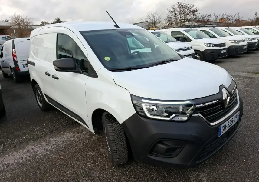 Renault Kangoo blanc en 3/4 avant droit sur parking gravillonné, avec calandre noire et phares allumés.