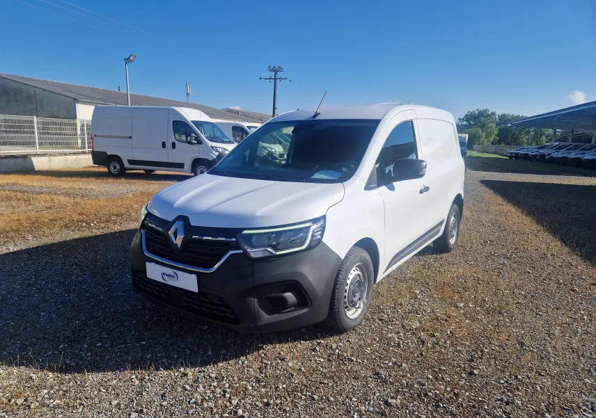 Renault Kangoo blanc en 3/4 avant droit, utilitaire avec pare-chocs noir et jantes acier, stationné sur sol gravillonné.