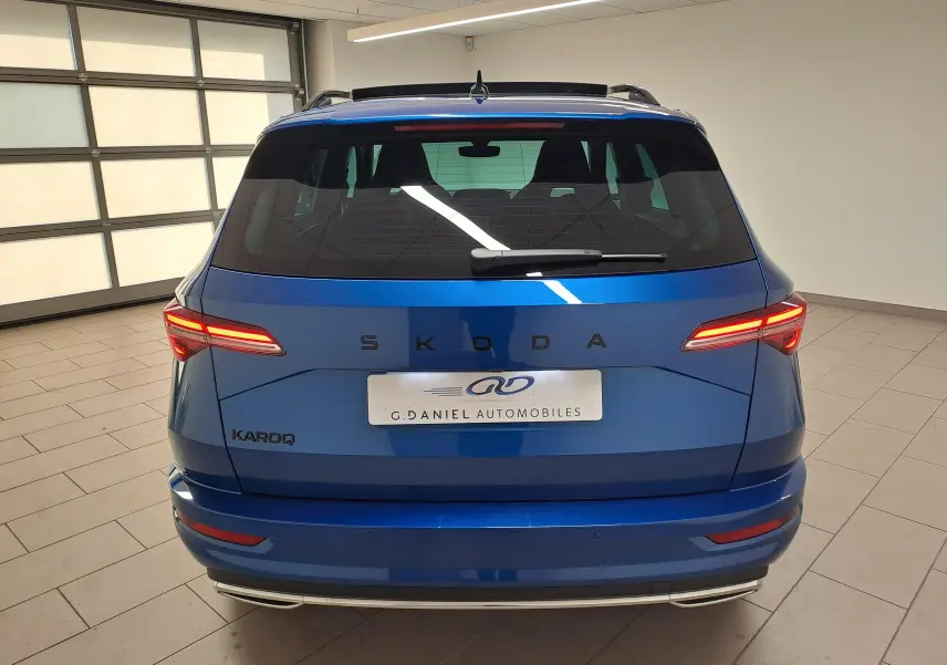 Vue arrière d'un Skoda Karoq Bleu RS 2025 avec feux allumés et toit panoramique ouvrant visible en intérieur showroom.