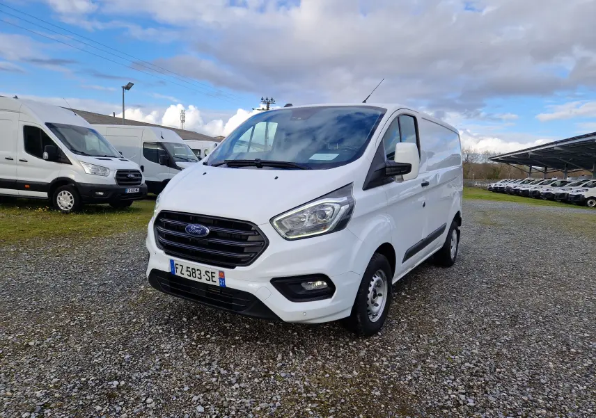Vue 3/4 avant droit d'un Ford Transit Custom fourgon blanc 2021 sur un parking extérieur avec plusieurs autres véhicules similaires.