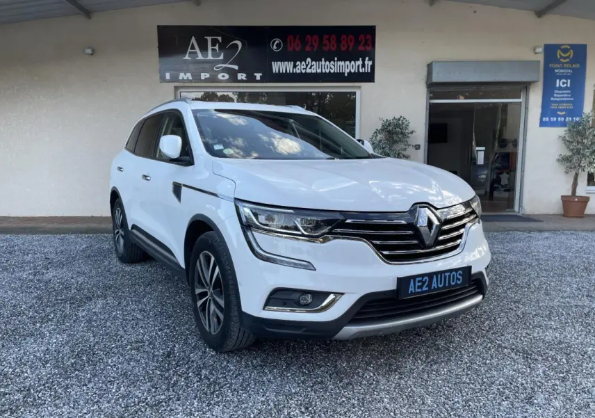 Renault Koleos blanc vu en 3/4 avant droit, avec calandre chromée et jantes alliage, stationné devant un bâtiment.