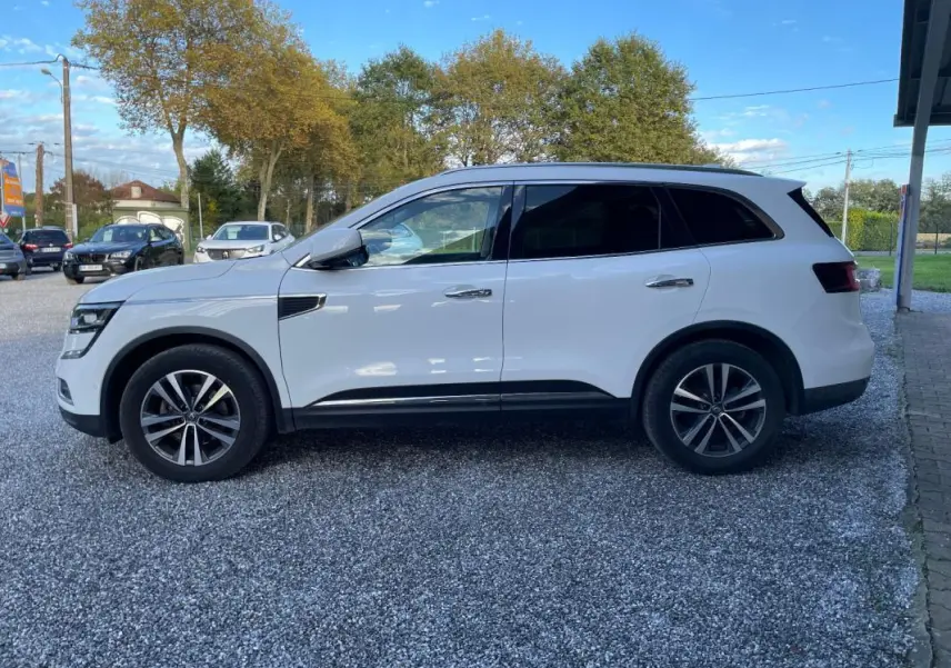 Renault Koleos blanc vu de profil côté gauche, SUV avec jantes alliage et vitres teintées, stationné sur gravier.