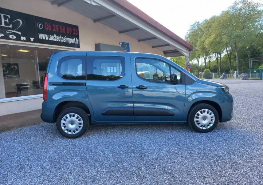 Vue de profil côté droit d'un Opel Combo Life Elegance M bleu 2025 avec protections latérales noires sur un sol gravillonné.