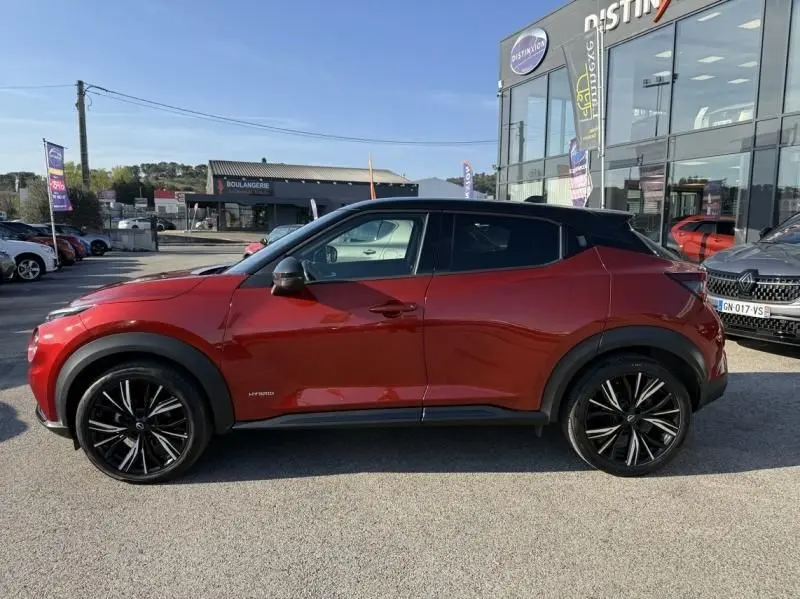 Vue latérale droite d'un Nissan Juke 1.6 Hybrid 143ch N-Design rouge Fuji métal avec toit noir, roues noires à jantes fines.