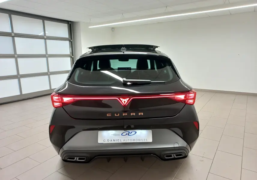 Vue arrière d'une CUPRA Leon 2025 Noir Minuit avec feux LED allumés et logo cuivré dans un garage blanc.