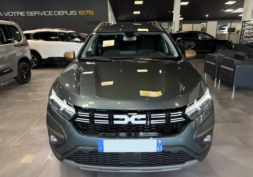 Vue frontale d'un Dacia Jogger Hybrid 140 Extreme vert cèdre en showroom avec calandre noire et phares allumés.