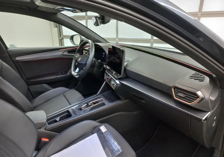Intérieur côté conducteur de la CUPRA Leon 2025, tableau de bord noir avec éclairage d'ambiance cuivré et écran tactile central.