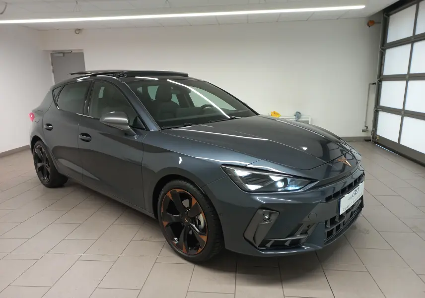 Vue 3/4 avant d'une CUPRA Leon 2025 gris magnétique avec jantes cuivrées et toit ouvrant panoramique en intérieur showroom.