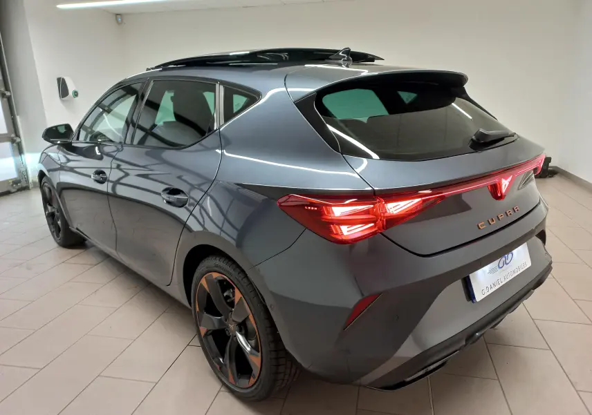 Vue 3/4 arrière droite d'une CUPRA Leon gris magnétique avec jantes cuivrées et toit ouvrant panoramique en intérieur.
