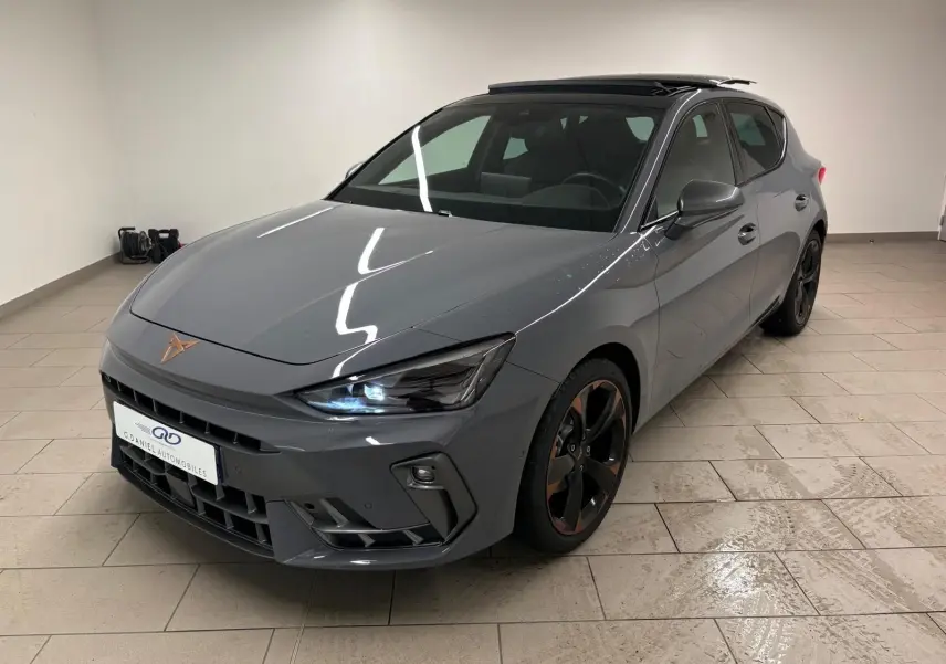Vue 3/4 avant droit d'une CUPRA Leon gris Graphène avec toit ouvrant panoramique et jantes alliage cuivrées en intérieur showroom.