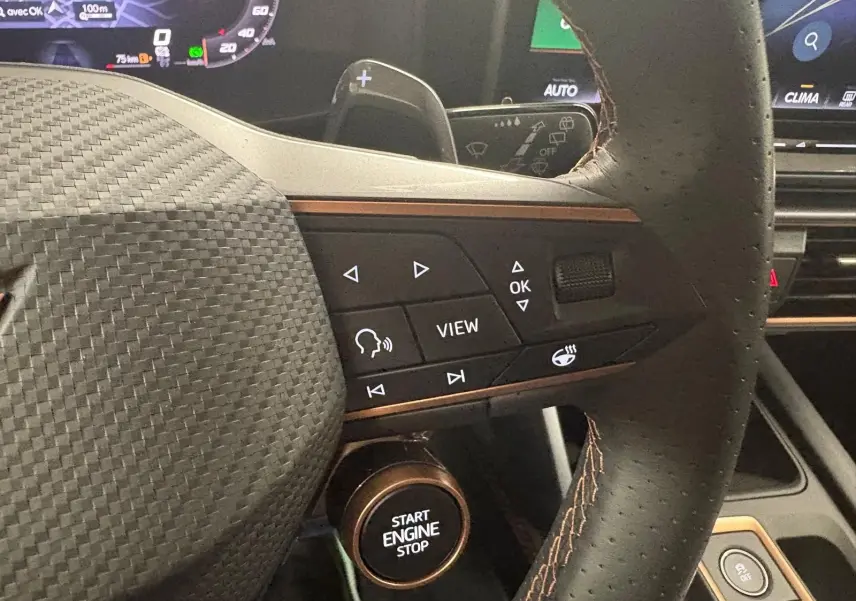 Gros plan sur le volant cuir noir avec surpiqûres cuivrées et commandes multifonctions du CUPRA Leon 2025, tableau de bord en arrière-plan.