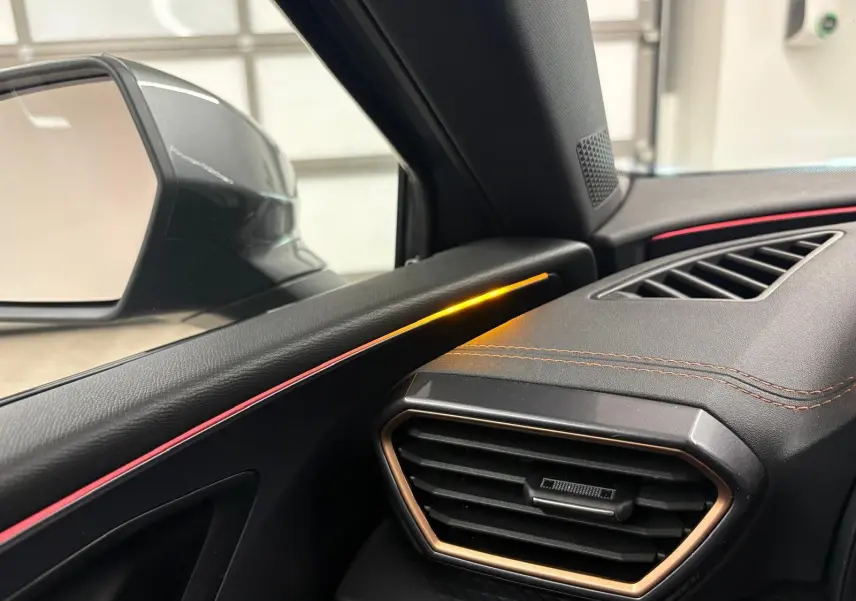 Détail intérieur côté gauche du tableau de bord de la CUPRA Leon 2025 avec éclairage d'ambiance orange et surpiqûres cuivrées.