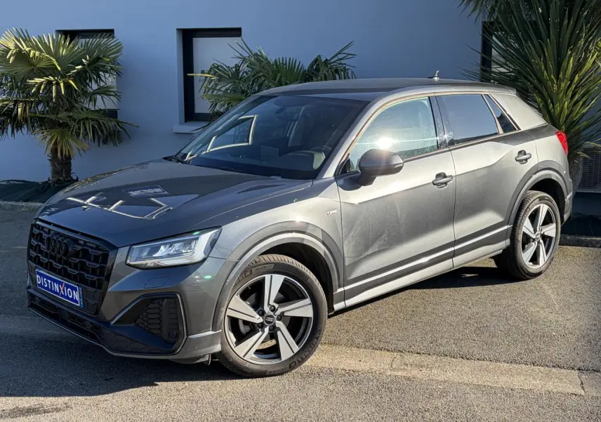 Audi Q2 35 TFSI S-Line gris Daytona en 3/4 avant droit, avec jantes alliage 18 pouces et vitres surteintées.
