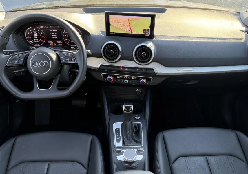 Vue intérieure avant de l'Audi Q2 35 TFSI S-Line 2024, tableau de bord noir avec écran GPS et volant multifonction cuir.