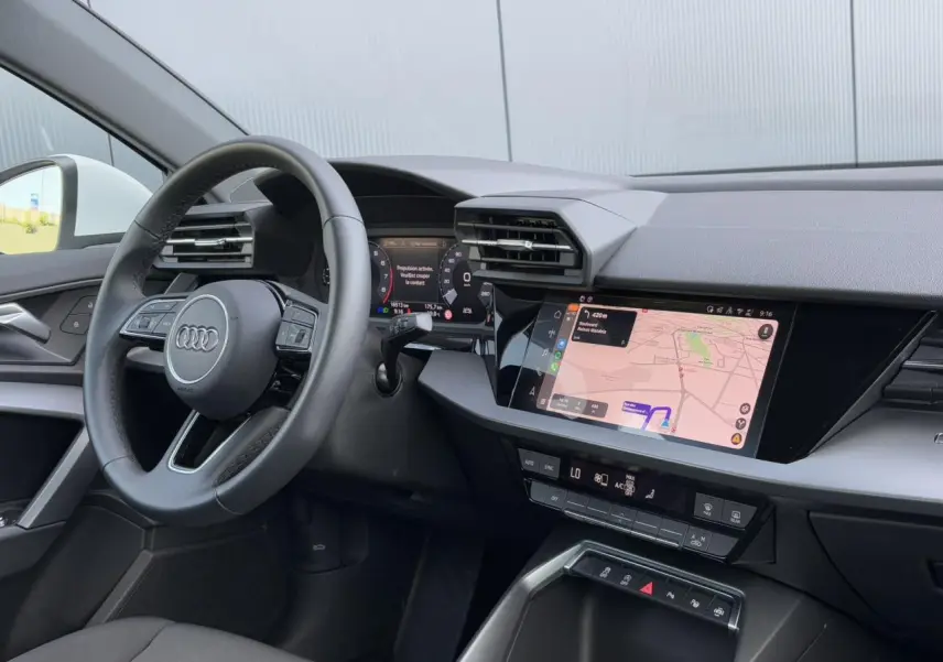 Vue intérieure côté conducteur de l'Audi A3 Sportback 2024, volant cuir et écran tactile de navigation allumé.