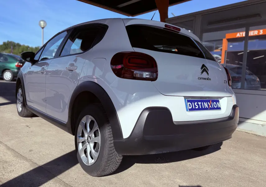 Citroën C3 blanc vue 3/4 arrière côté gauche, avec feux arrière sombres et protections noires sur les ailes.