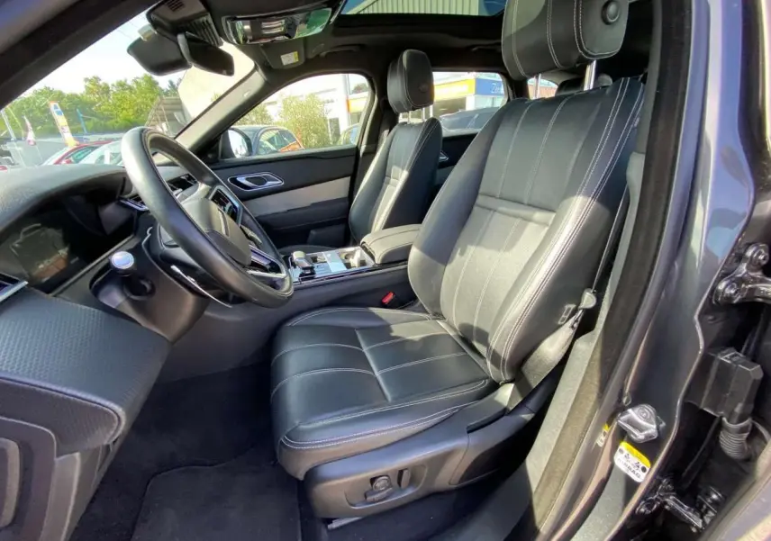 Vue intérieure côté conducteur du Range Rover Velar gris tempête, sièges cuir noirs avec surpiqûres blanches et console centrale moderne.