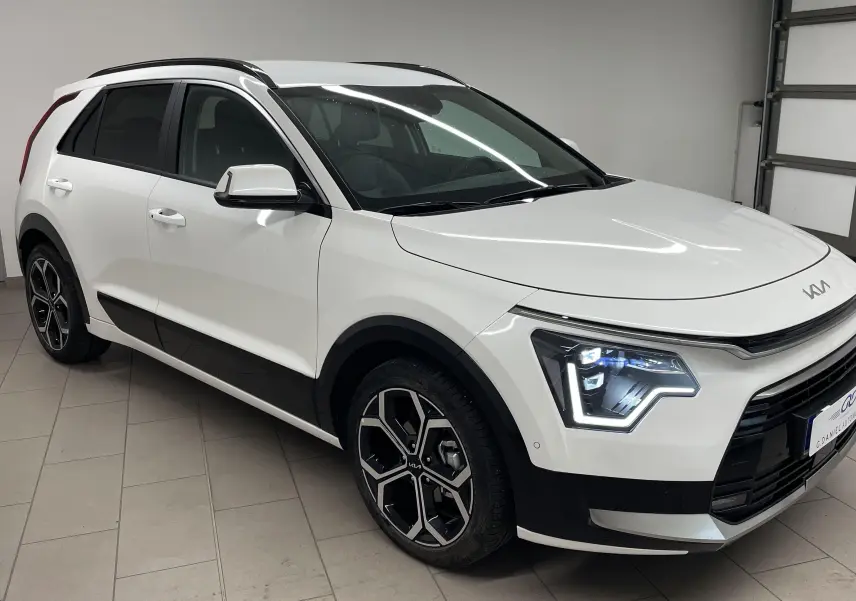 KIA NIRO blanc 2025 en 3/4 avant droit, avec phares LED distinctifs et jantes alliage noires et argentées.