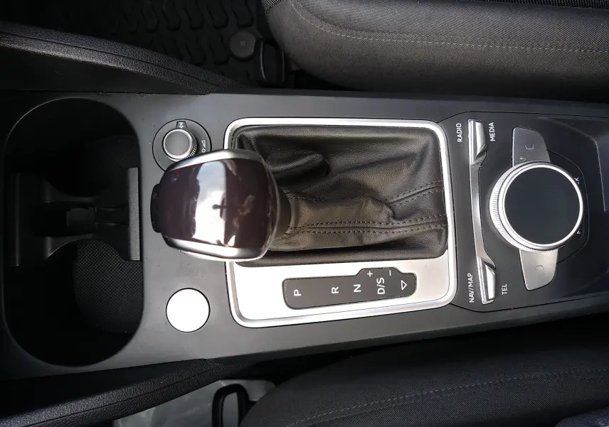 Vue rapprochée de la console centrale de l'Audi Q2 noire, mettant en valeur le levier de vitesses automatique et la molette de contrôle multimédia.