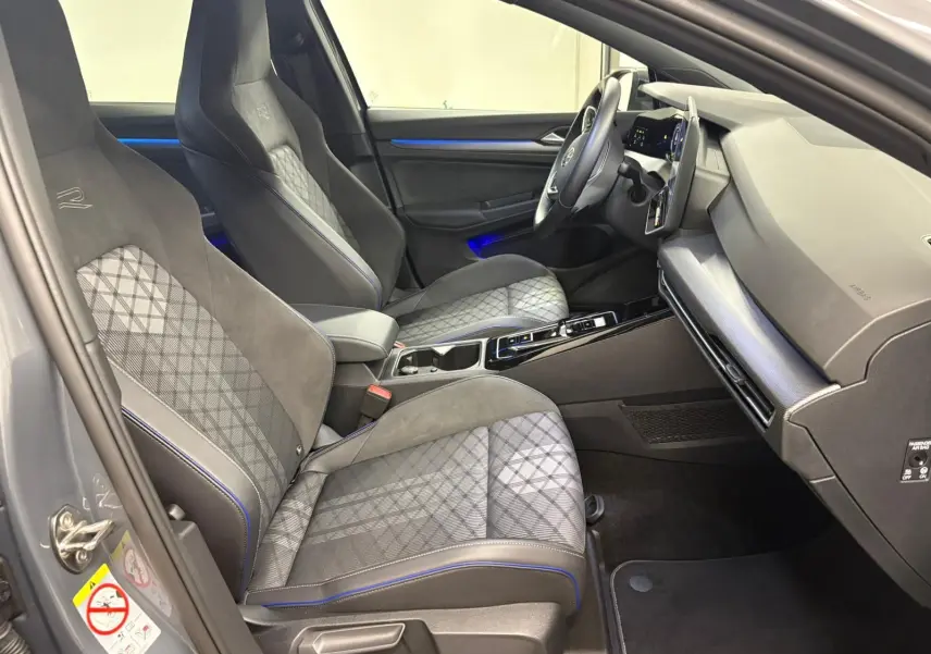 Vue intérieure côté gauche de la Volkswagen Golf 1.5 eTSI R-Line 2025 avec sièges sport gris à surpiqûres bleues et tableau de bord moderne.