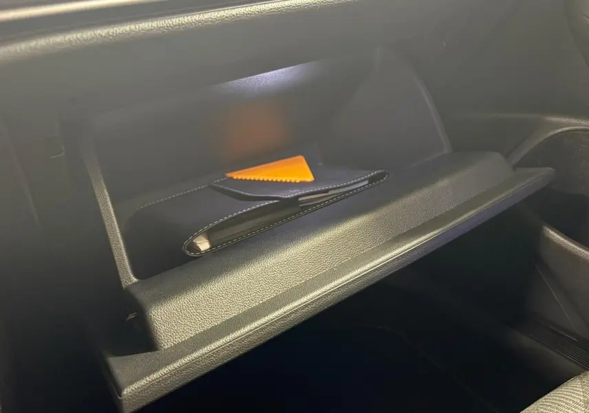 Gros plan sur la boîte à gants ouverte de la Volkswagen Golf 1.5 eTSI 150 DSG7 R-Line Edition avec un livret et un grattoir orange à l'intérieur.