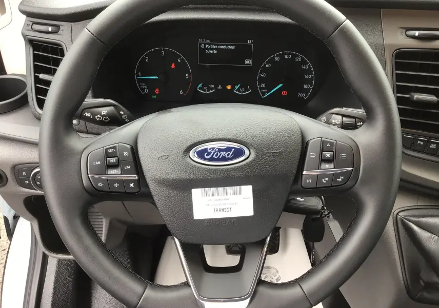 Vue rapprochée du volant noir du Ford Transit blanc 2023 avec commandes intégrées et tableau de bord numérique.