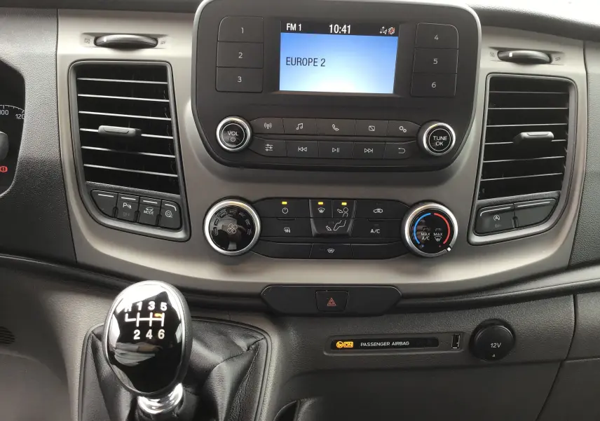 Tableau de bord du Ford Transit Fourgon 2023 avec écran radio, commandes climatisation et levier de vitesse manuel en vue frontale.