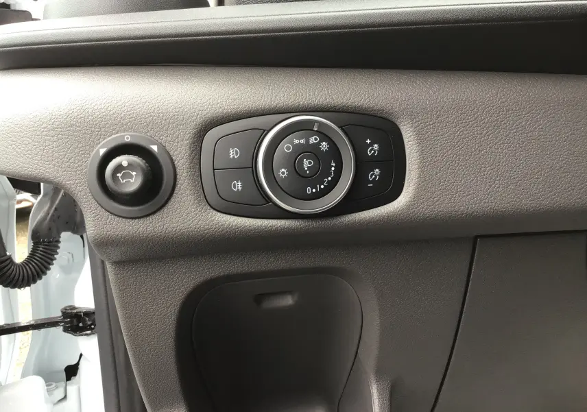 Gros plan sur les commandes d'éclairage et réglage des phares du Ford Transit Fourgon blanc, côté gauche du tableau de bord.