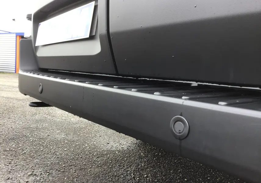 Gros plan sur le pare-chocs arrière noir du Ford Transit Fourgon blanc, mettant en valeur les capteurs de recul intégrés.