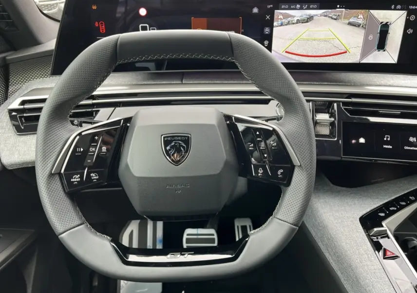 Volant cuir gris avec logo Peugeot et inscription GT, tableau de bord numérique affichant caméra de recul, intérieur Alcantara gris titane.