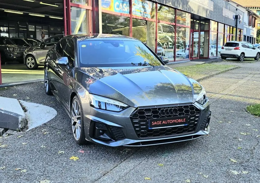 Audi A5 Sportback gris Daytona vue 3/4 avant, avec calandre noire hexagonale et jantes alliage visibles devant un garage.