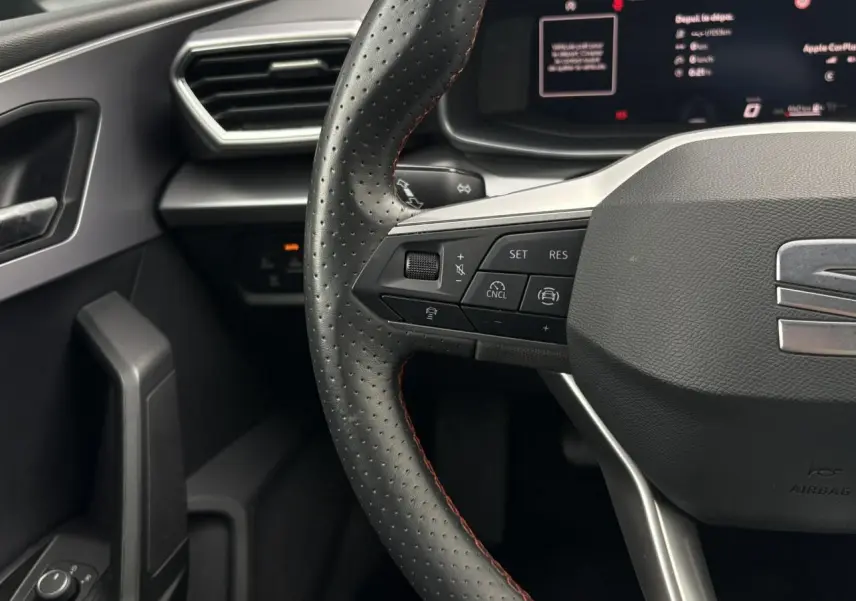 Gros plan sur le volant en cuir noir perforé avec surpiqûres rouges du SEAT Leon 2025, tableau de bord numérique en arrière-plan.