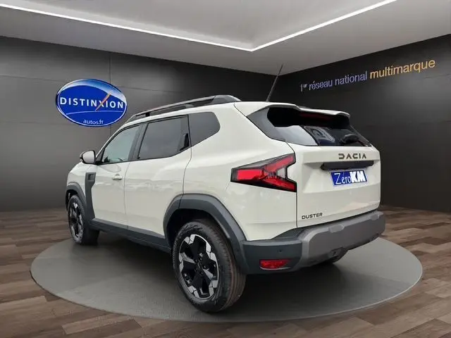 Vue 3/4 arrière droite d'un Dacia Duster blanc avec jantes alu noires et toit noir dans un showroom.