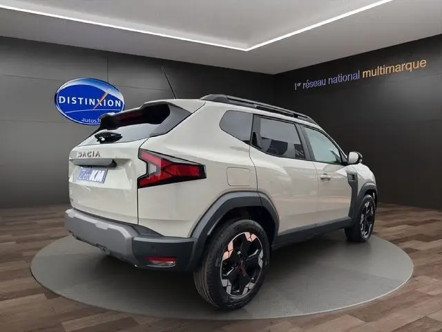 Vue 3/4 arrière droite d'un Dacia Duster blanc avec jantes alu noires et barres de toit en showroom.