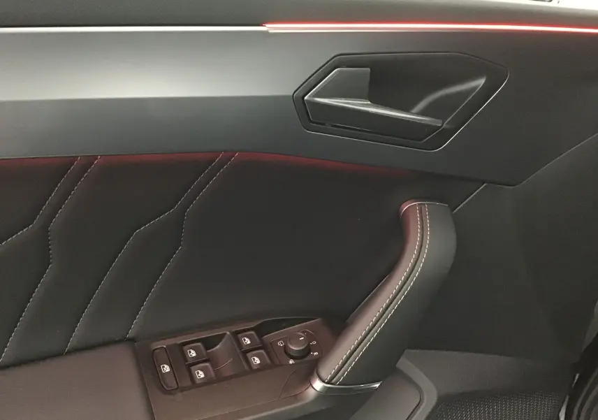 Intérieur côté gauche de la porte d'une CUPRA Leon noire 2023, avec surpiqûres blanches et commandes de vitres intégrées.