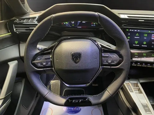 Vue rapprochée du volant noir perforé de la Peugeot 308 GT hybride 2026 avec tableau de bord digital et écran tactile.