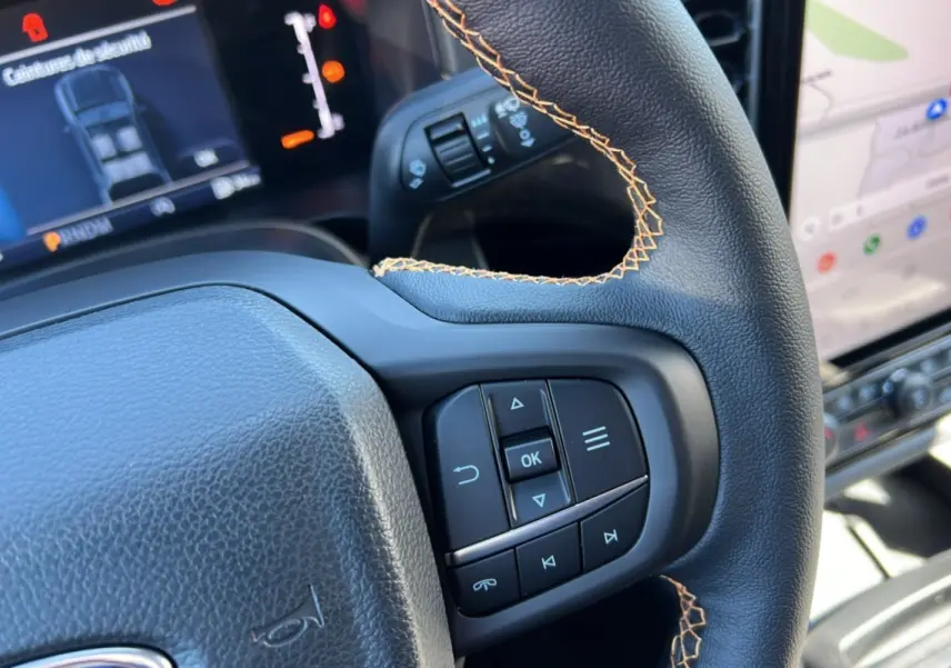 Gros plan sur le volant avec surpiqûres orange du Ford Ranger 2025 gris carbone, montrant les commandes intégrées et l'écran digital.