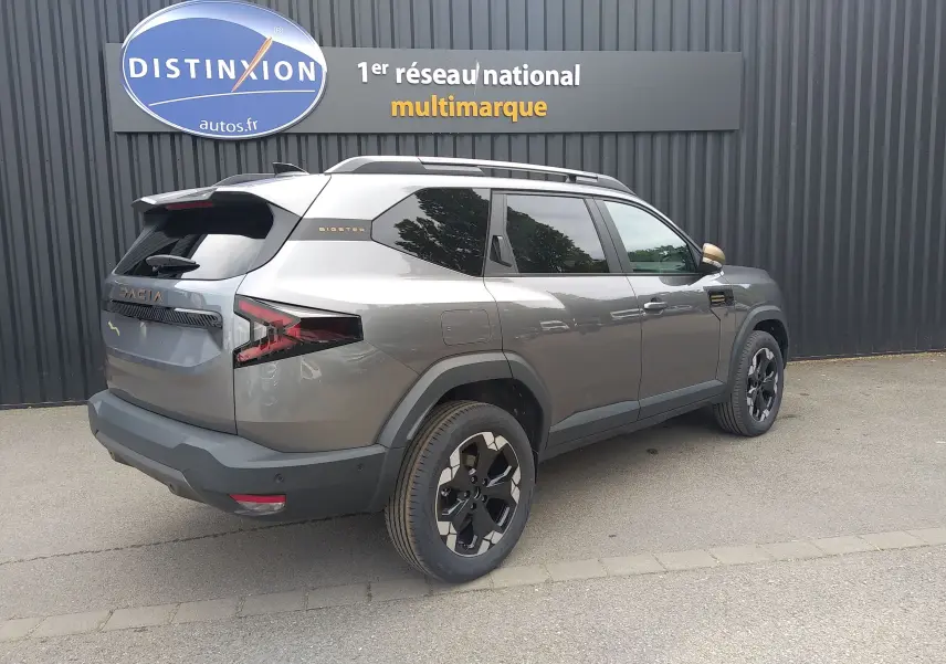 Vue 3/4 arrière droite du Dacia Bigster gris schiste avec jantes noires et détails bronze, stationné devant un mur sombre.