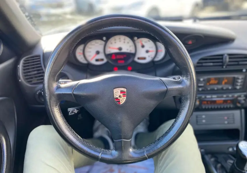 Vue intérieure centrée sur le volant en cuir noir avec logo Porsche, tableau de bord blanc et levier de vitesse manuel visible.
