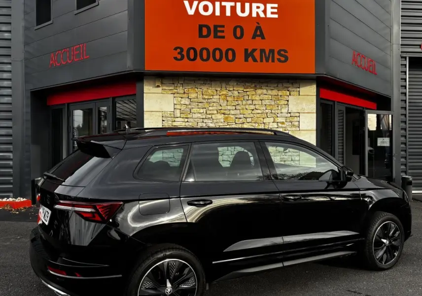 Vue 3/4 arrière droite d'un SUV SKODA KAROQ noir brillant garé devant un bâtiment avec une enseigne orange.