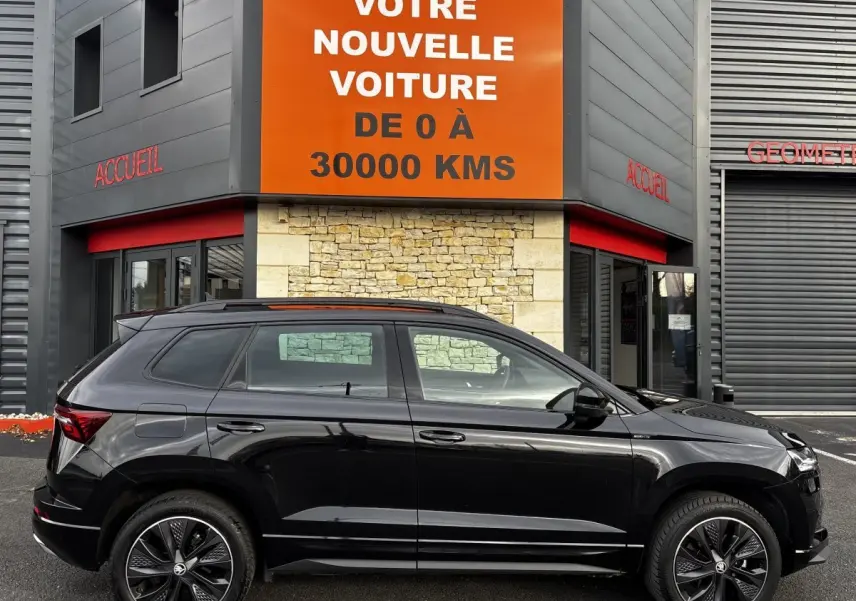 Profil côté gauche d'un Skoda Karoq noir 2025 garé devant un bâtiment avec panneau orange "Votre nouvelle voiture".