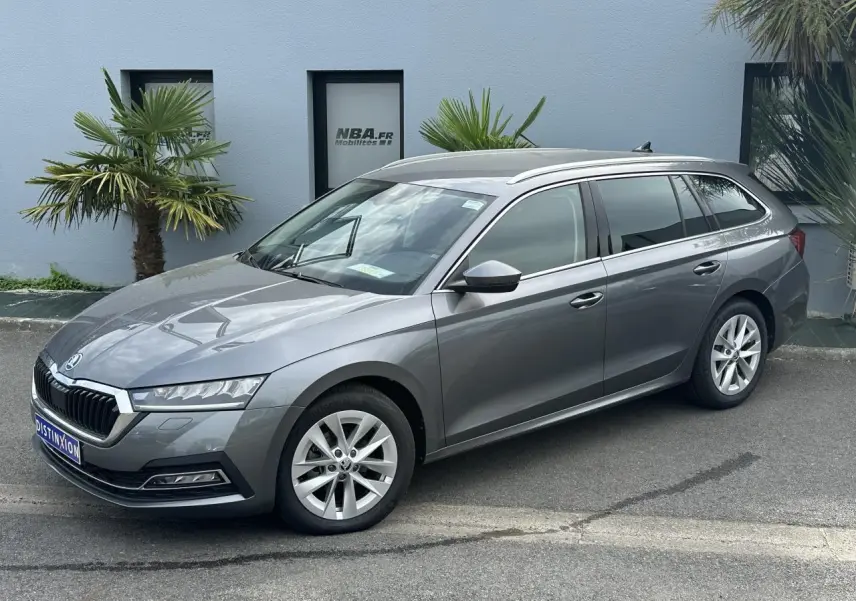 Skoda Octavia Combi gris graphite vue 3/4 avant droit, avec jantes alliage et vitres teintées, stationnée devant un mur bleu.
