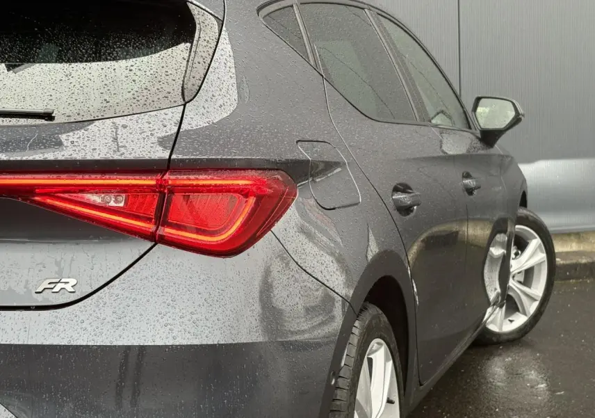 Vue 3/4 arrière droite d'une SEAT LEON 1.5 e-TSI FR gris magnétique avec feux arrière LED allumés sous la pluie.